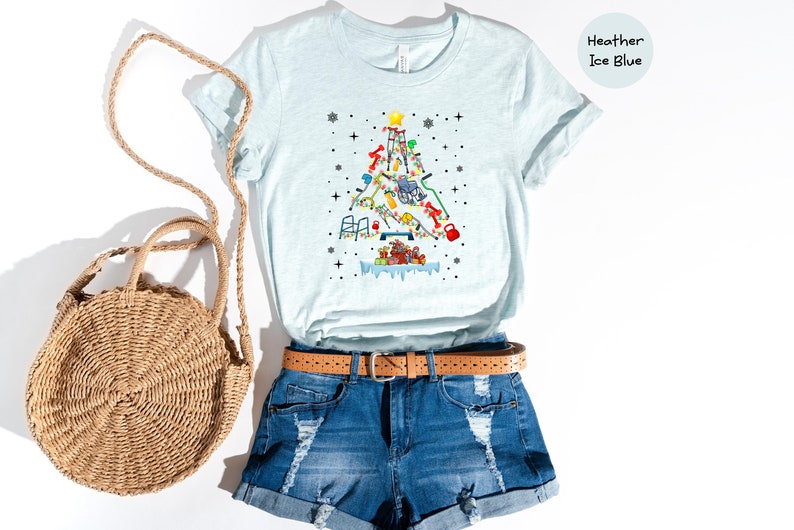 May include: A light blue t-shirt with a Christmas tree design made up of various fitness equipment. The tree is decorated with lights and presents. The shirt is displayed on a model with denim shorts and a straw bag.