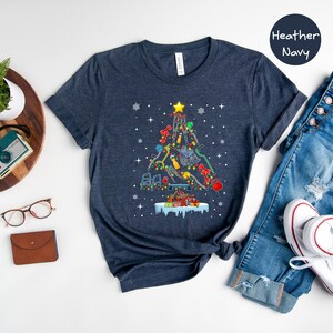 May include: A heather navy blue t-shirt with a Christmas tree design made up of fitness equipment. The tree is decorated with lights and ornaments. The shirt is laid flat on a white surface with a pair of jeans, a pair of white sneakers, a potted plant, a watch, a camera, and a pair of glasses.