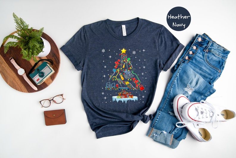 May include: A heather navy blue t-shirt with a Christmas tree design made up of fitness equipment. The tree is decorated with lights and ornaments. The shirt is laid flat on a white surface with a pair of jeans, a pair of white sneakers, a potted plant, a watch, a camera, and a pair of glasses.