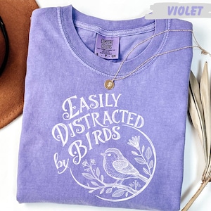 a purple t-shirt with a white graphic design, a necklace, a hat, and a plant.