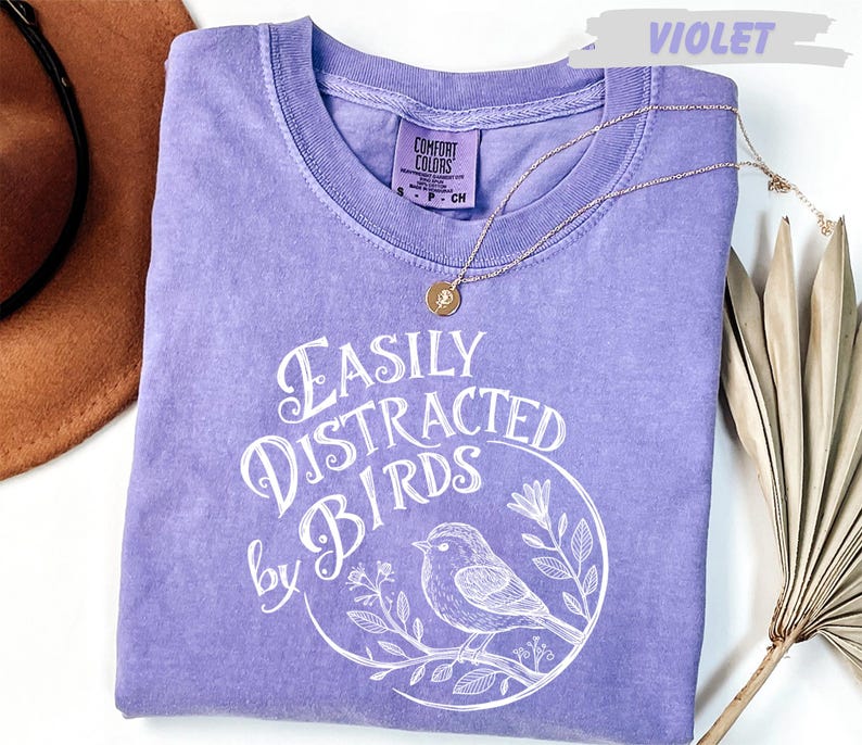 a purple t-shirt with a white graphic design, a necklace, a hat, and a plant.