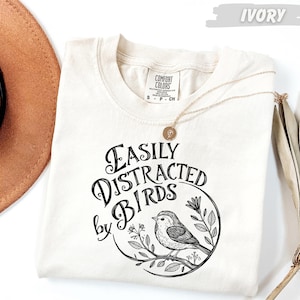 a white t-shirt with a black and white graphic design that says "Easily Distracted by Birds" and includes a small bird illustration. The t-shirt is laid out on a white surface, with a brown hat and a fan visible in the background.