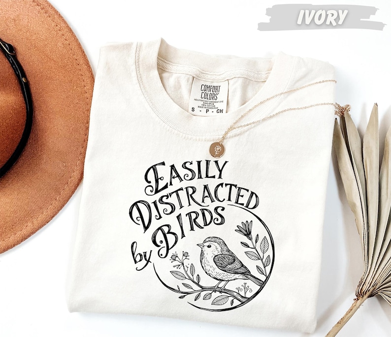a white t-shirt with a black and white graphic design that says "Easily Distracted by Birds" and includes a small bird illustration. The t-shirt is laid out on a white surface, with a brown hat and a fan visible in the background.
