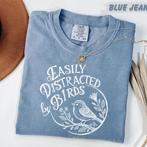 a blue t-shirt with a white graphic design, a brown hat, and some dried leaves, all arranged on a white surface.