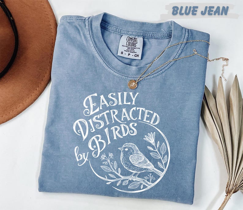 a blue t-shirt with a white graphic design, a brown hat, and some dried leaves, all arranged on a white surface.