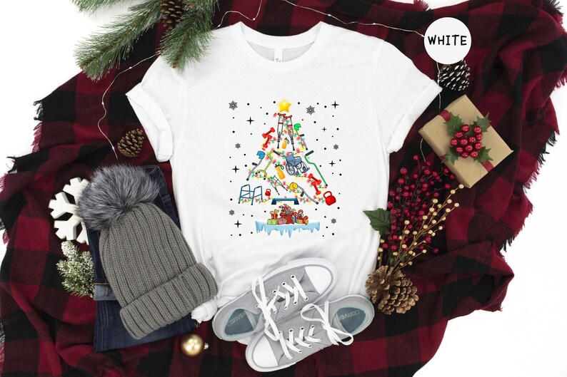 May include: White t-shirt with a Christmas tree design made up of medical equipment, including crutches, a walker, a wheelchair, and a weight. The tree is decorated with lights and ornaments. The t-shirt is on a red and black plaid blanket with a gray beanie, a pair of gray sneakers, and a small pine cone.