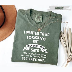 May include: A moss green short-sleeved t-shirt featuring white text that reads: I WANTED TO GO JOGGING BUT PROVERBS 28:1 SAYS The Wicked Run When No One Is Chasing Them SO THERE'S THAT... The shirt has a crew neckline and a Comfort Colors brand label.