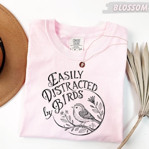 a pink t-shirt with a graphic design of a bird and the text "Fasily Distracted by Birds" printed on it. The t-shirt is laid out on a white surface, along with a brown hat and a fan.