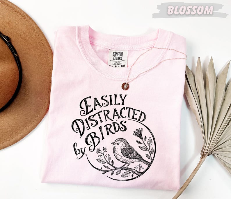 a pink t-shirt with a graphic design of a bird and the text "Fasily Distracted by Birds" printed on it. The t-shirt is laid out on a white surface, along with a brown hat and a fan.