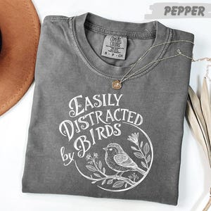 a gray t-shirt with a white graphic design, a brown hat, and some dried leaves, all placed on a white surface.