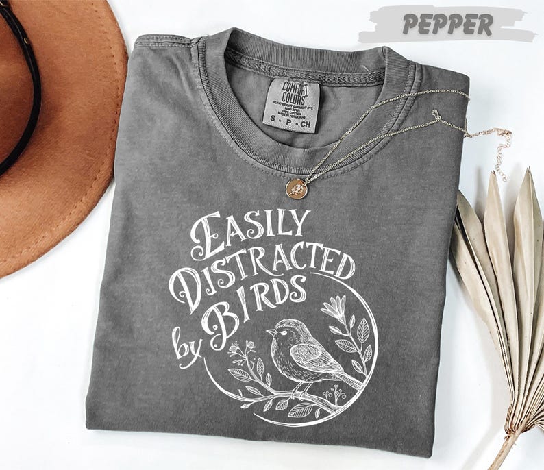 a gray t-shirt with a white graphic design, a brown hat, and some dried leaves, all placed on a white surface.