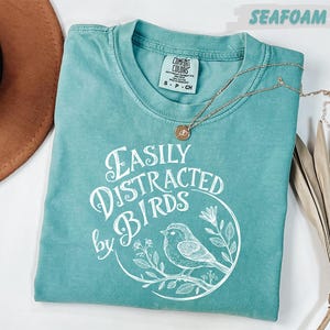 a turquoise t-shirt with a white design of a bird and the text "Easily Distracted by Birds" printed on it. The t-shirt is laid out on a white surface, accompanied by a brown hat, a necklace, and some dried leaves.