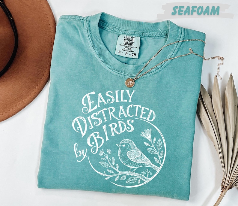 a turquoise t-shirt with a white design of a bird and the text "Easily Distracted by Birds" printed on it. The t-shirt is laid out on a white surface, accompanied by a brown hat, a necklace, and some dried leaves.