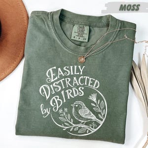 May include: A moss green t-shirt with the text "Easily Distracted by Birds" in white script, featuring a bird illustration. A gold necklace with a small pendant rests on the shirt. The shirt is made of a soft material.