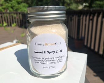 May include: A clear glass jar with a silver lid filled with a light-colored powder. The jar has a white label with the text "Saucy Brown Girl Sweet & Spicy Chai." The label lists ingredients and the net weight of 2.5 oz (71 g).