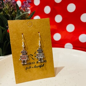 May include: A pair of silver robot earrings on a brown card with the text "BOOBOO & MOO Bold is beautiful".