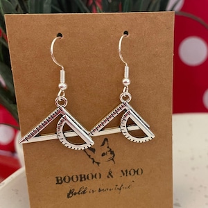 Puede incluir: Pendientes de plata con diseño de transportador y triángulo. Los pendientes se muestran en una tarjeta marrón con el texto "Booboo & Moo Bold is beautiful."