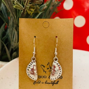 May include: Silver-coloured dangle earrings in the shape of pierogies, with detailed textures. The earrings hang from silver hooks and are displayed on a brown card with the text "BOO & CO" and "Bold is beautiful". The background includes greenery and a red polka-dot pattern.