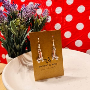 May include: A pair of silver-toned roller skate earrings displayed on a rectangular brown card. The card features the text BOOBOO & MOO with a small line drawing of a dog above it. The earrings have a metallic finish and hook-style backings.