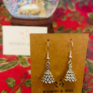 Puede incluir: Un par de pendientes plateados con forma de árbol de Navidad y detalles de estrella, expuestos en una tarjeta marrón. Los pendientes están delante de un fondo borroso con una bola de nieve y una tarjeta con el texto "BOOBOO & MOO".