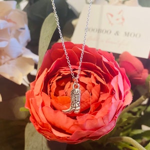 May include: A silver-colored necklace with a small cowboy boot pendant. The necklace is displayed against a backdrop of a large, vibrant red rose. The chain is delicate and the boot pendant has intricate detailing.