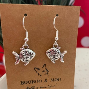 Può includere: Orecchini a forma di pesce color argento con squame e pinne dettagliate, appesi a ganci d'argento. Gli orecchini sono esposti su una carta marrone con il testo "BOOBOO & MOO" e "Bold is beautiful."