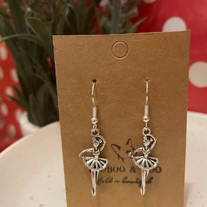 May include: Silver-toned ballerina earrings with a tutu and arms raised. The earrings hang from silver hooks and are displayed on a brown card. The card has the text "BOO & CO" and "Bold is beauty."