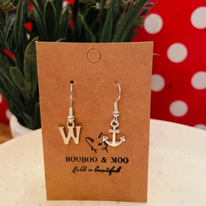 May include: Silver-toned dangle earrings displayed on a brown card. One earring features the letter "W", the other an anchor charm. The card has the text "BOOBOO & MOO" and "bold is beautiful". The background includes greenery and a red polka-dot pattern.