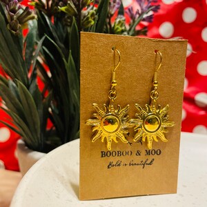 Op de afbeelding: Goudkleurige zonne-oorbellen met een haaksluiting. De oorbellen worden gepresenteerd op een bruine kaart met de tekst "BOOBOO & MOO" en "Bold is beautiful". De achtergrond bevat groen en een rood-wit stippenpatroon.