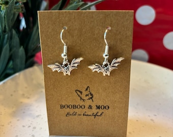 May include: A pair of silver-toned bat-shaped dangle earrings with hook wires, displayed on a rectangular brown card. The card features a line drawing of a dog, the text BOOBOO & MOO, and the phrase Bold is beautiful printed in black ink.