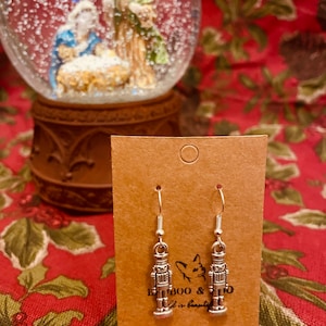 Può includere: Orecchini a forma di schiaccianoci in metallo color argento su una carta marrone. Gli orecchini sono appesi a un gancio d'argento. La carta ha il testo "BOO & ... is beautiful". Una palla di neve con una scena della natività è sullo sfondo.