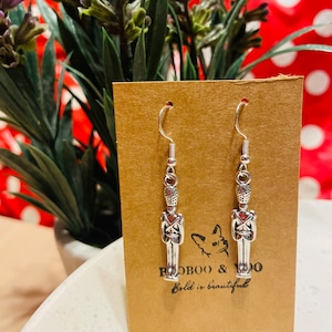 Può includere: Un paio di orecchini color argento a forma di soldatini, appesi a ganci d'argento. Gli orecchini sono esposti su una carta marrone con il testo "BOOBOO & DOO Bold is beautiful". Lo sfondo include una pianta con fiori viola e un motivo a pois rossi e bianchi.