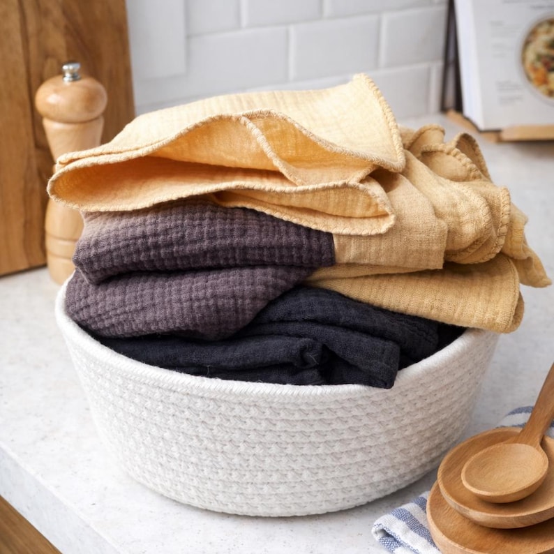May include: A white woven basket filled with folded kitchen towels in shades of yellow, purple, and dark gray. The towels are stacked inside the basket, creating a layered effect. Wooden spoons and a pepper grinder are in the background.