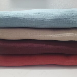 May include: A stack of four folded, textured fabric cloths in shades of blue, beige, burgundy, and coral. The cloths are neatly stacked, showcasing the different colors and textures of the fabric. The image highlights the soft, layered appearance of the textiles.
