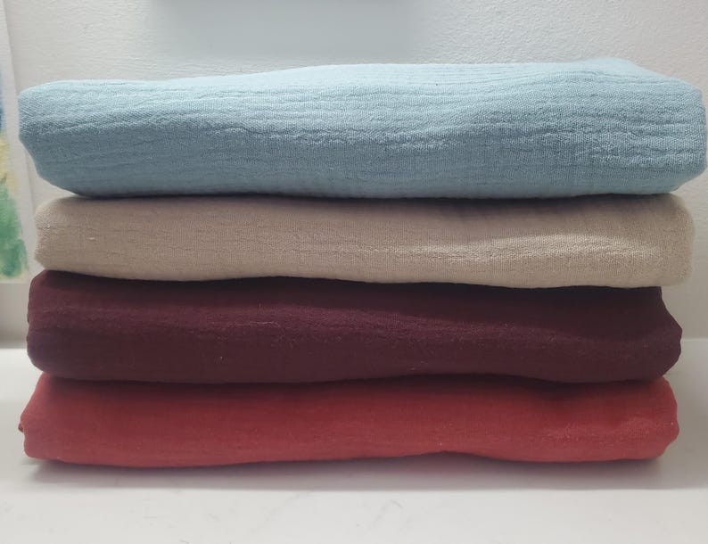 May include: A stack of four folded, textured fabric cloths in shades of blue, beige, burgundy, and coral. The cloths are neatly stacked, showcasing the different colors and textures of the fabric. The image highlights the soft, layered appearance of the textiles.
