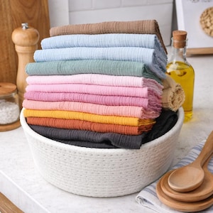 May include: A stack of folded, textured fabric cloths in various colors including brown, light blue, sage green, pink, yellow, orange, and black, arranged inside a round, white, woven rope basket with visible stitching details.