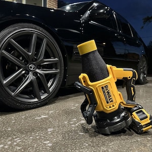 Peut inclure: Un souffleur de feuilles sans fil DeWalt jaune et noir avec une buse en mousse noire. Le souffleur est posé sur une surface en béton gris devant une voiture noire.