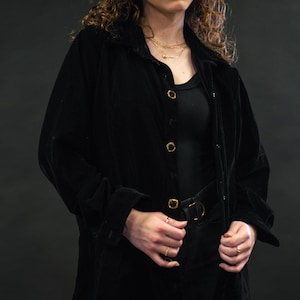 May include: A woman wearing a black velvet shirt with gold buttons, a black tank top, black jeans, and gold jewelry. She is standing in front of a dark background.