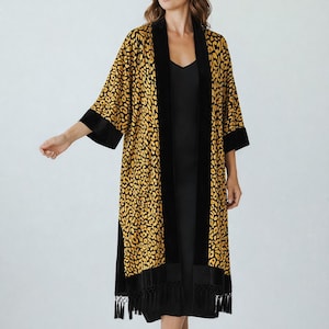 May include: A long, open-front robe with a yellow and black leopard print. The robe has black velvet trim on the collar, cuffs, and down the front. The hemline features black fringe. The robe is worn over a black dress.
