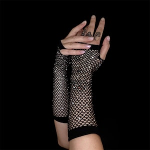 May include: Black fishnet gloves with sparkling rhinestones. The gloves are fingerless, extending from the wrist to the mid-forearm. The background is solid black, highlighting the texture and sparkle of the gloves.