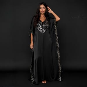 May include: A floor-length black kaftan dress featuring a V-neckline and wide sleeves. The garment is adorned with a decorative panel of reflective silver-toned metallic accents around the neckline and along the vertical edges of the sleeves.