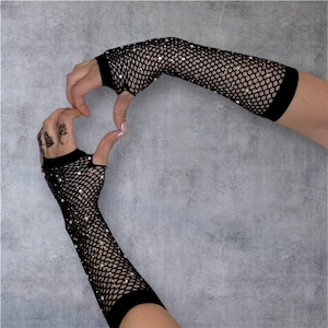 May include: Black fishnet gloves with sparkling embellishments. The gloves are fingerless, extending up the arm to the elbow. The hands are positioned to form a heart shape against a gray background.