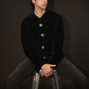 May include: A man wearing a black velvet shirt with gold buttons sits on a metal stool. He is wearing black jeans and black shoes.