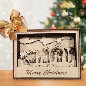 Può includere: Decorazione natalizia in legno con un presepe tagliato al laser, palme, cammelli e una stella. Le parole "Merry Christmas" sono in basso. Un fiocco dorato è nell'angolo in alto a sinistra.