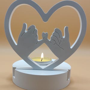 Può includere: Portacandele in legno bianco a forma di cuore con due mani che formano una promessa mignolo all'interno del cuore. Il portacandele ha una piccola base rotonda e una candela è posizionata al centro.