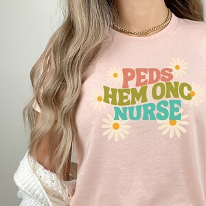 May include: A heather peach t-shirt with the text "PEDS HEM ONC NURSE" in colorful letters and daisy flowers around the text.