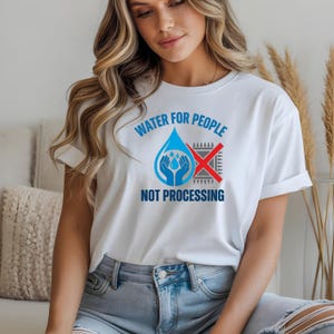 May include: A white short-sleeved t-shirt featuring a graphic design with the text WATER FOR PEOPLE NOT PROCESSING. The graphic includes a blue water droplet icon containing two hands holding water, next to a grey computer chip with a red X over it.