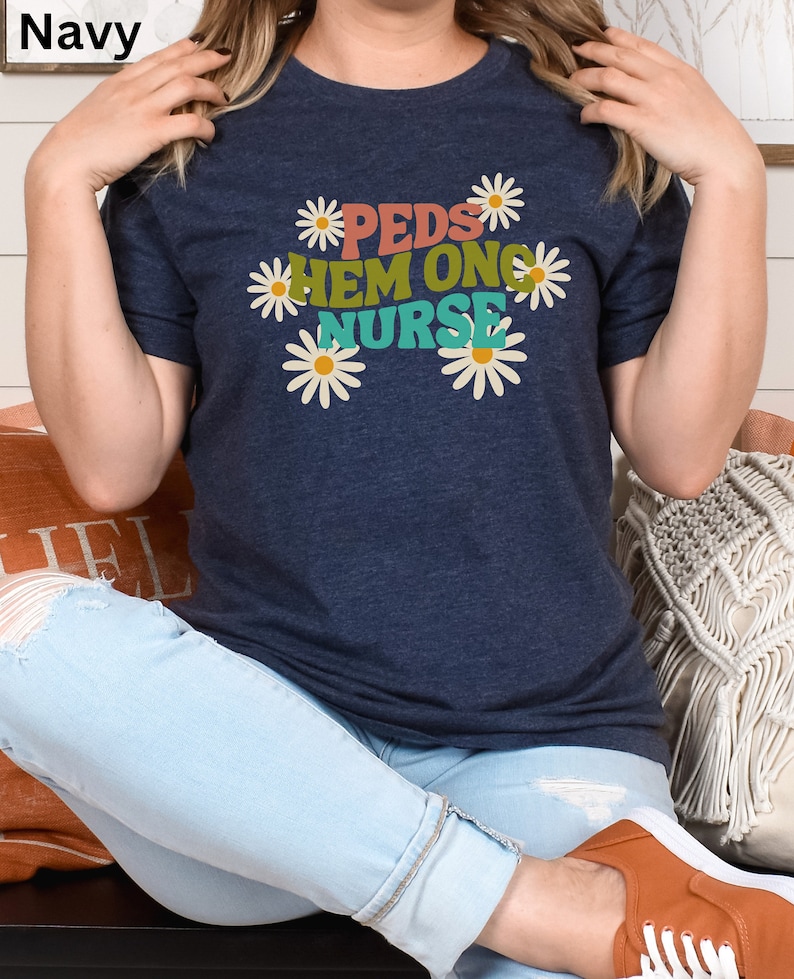 May include: A navy blue t-shirt with a graphic design of daisies and the text "PEDS HEM ONC NURSE" in colorful letters.