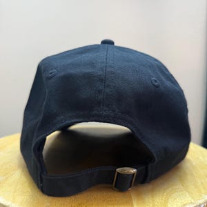 K&ouml;nnte beinhalten: Eine schwarze Baseballkappe mit gebogenem Schirm und verstellbarem Riemen. Die Kappe besteht aus einem einfarbigen, dunklen Stoff und hat einen Metall-Schnallenverschluss. Die Kappe liegt auf einer Holzoberfl&auml;che.
