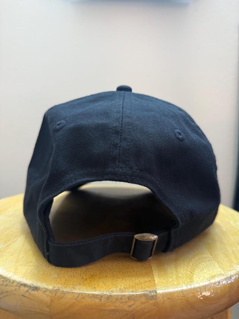 K&ouml;nnte beinhalten: Eine schwarze Baseballkappe mit gebogenem Schirm und verstellbarem Riemen. Die Kappe besteht aus einem einfarbigen, dunklen Stoff und hat einen Metall-Schnallenverschluss. Die Kappe liegt auf einer Holzoberfl&auml;che.